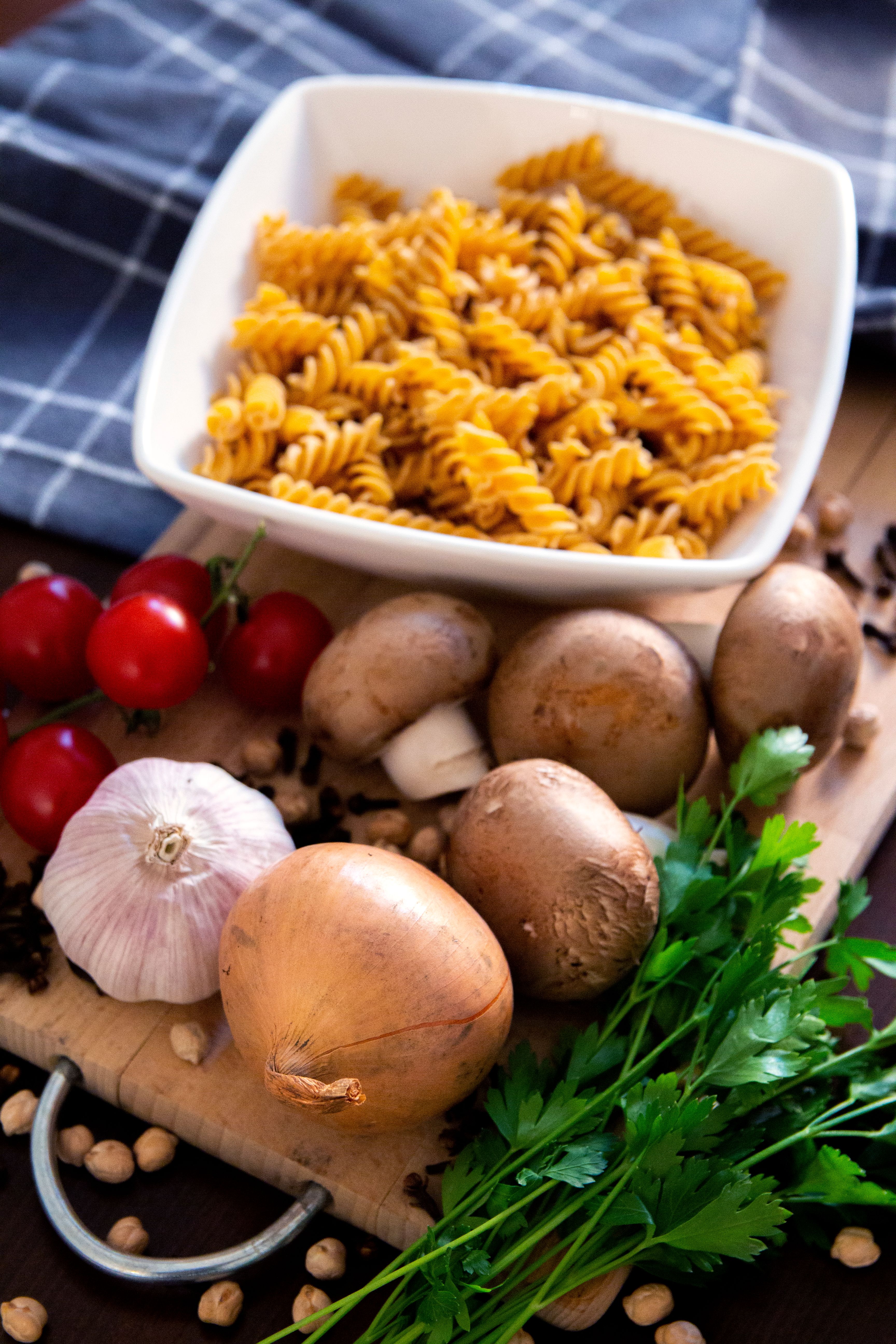 Bowl of fusilli pasta with garlic, onion, mushrooms, tomatoes, and parsley on a wooden board.