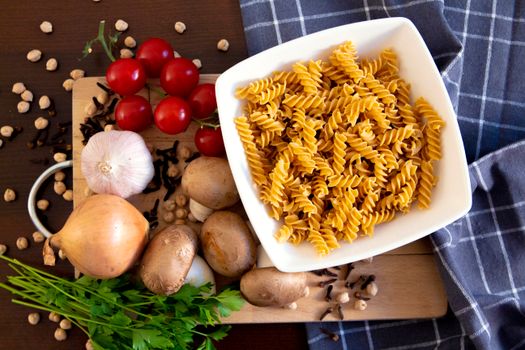 Top view of uncooked fusilli pasta with garlic, tomatoes, mushrooms, onions, and herbs on a wooden board.