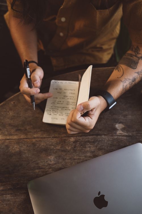 Free Man with tattoos writing in a notebook beside a laptop on a wooden table, Bali. Stock Photo