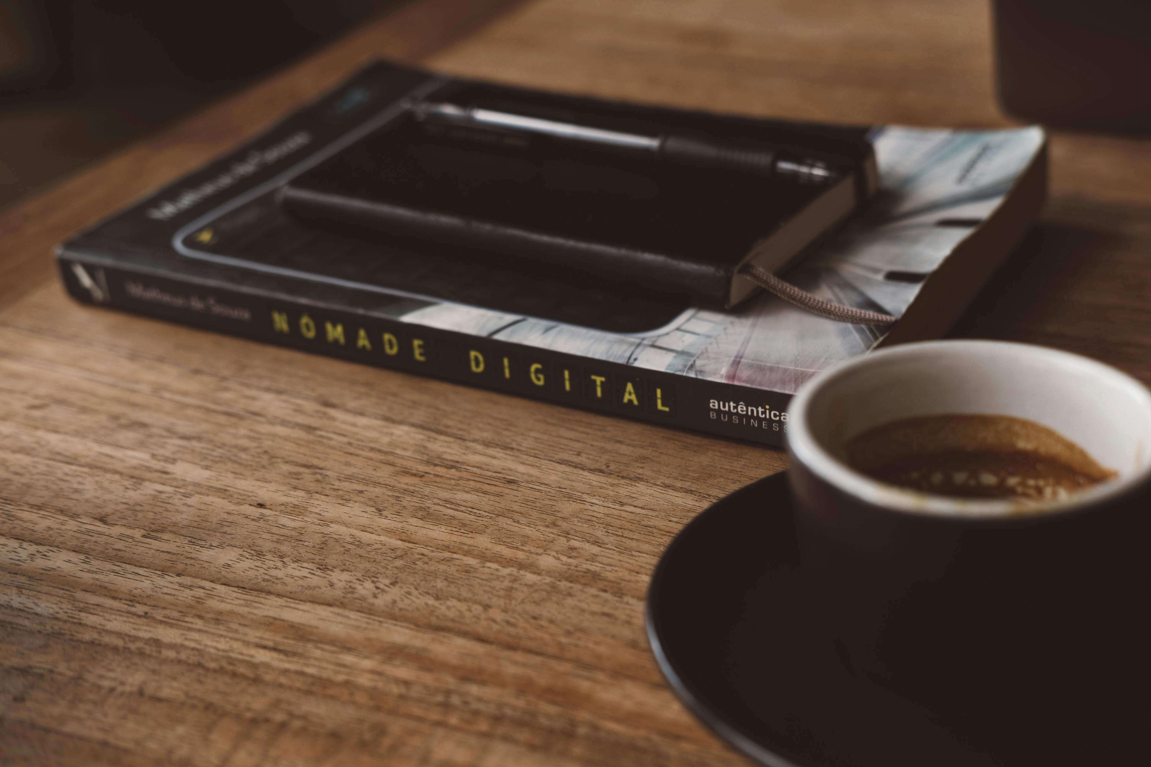 Close-up of a book, notebook, and coffee cup on a wooden table, perfect for a work-from-cafe theme.