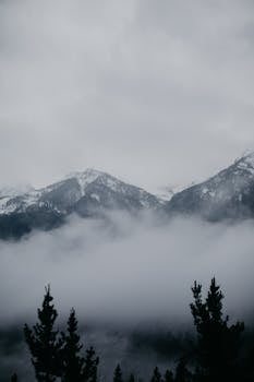 A dramatic, foggy mountain landscape showcasing snow-covered peaks under an overcast sky.