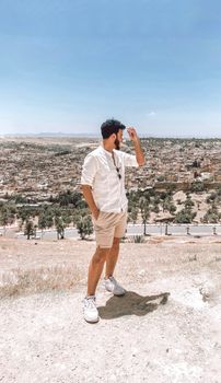 Man standing stylishly with a cityscape of Fes, Morocco in the background under a clear sky.
