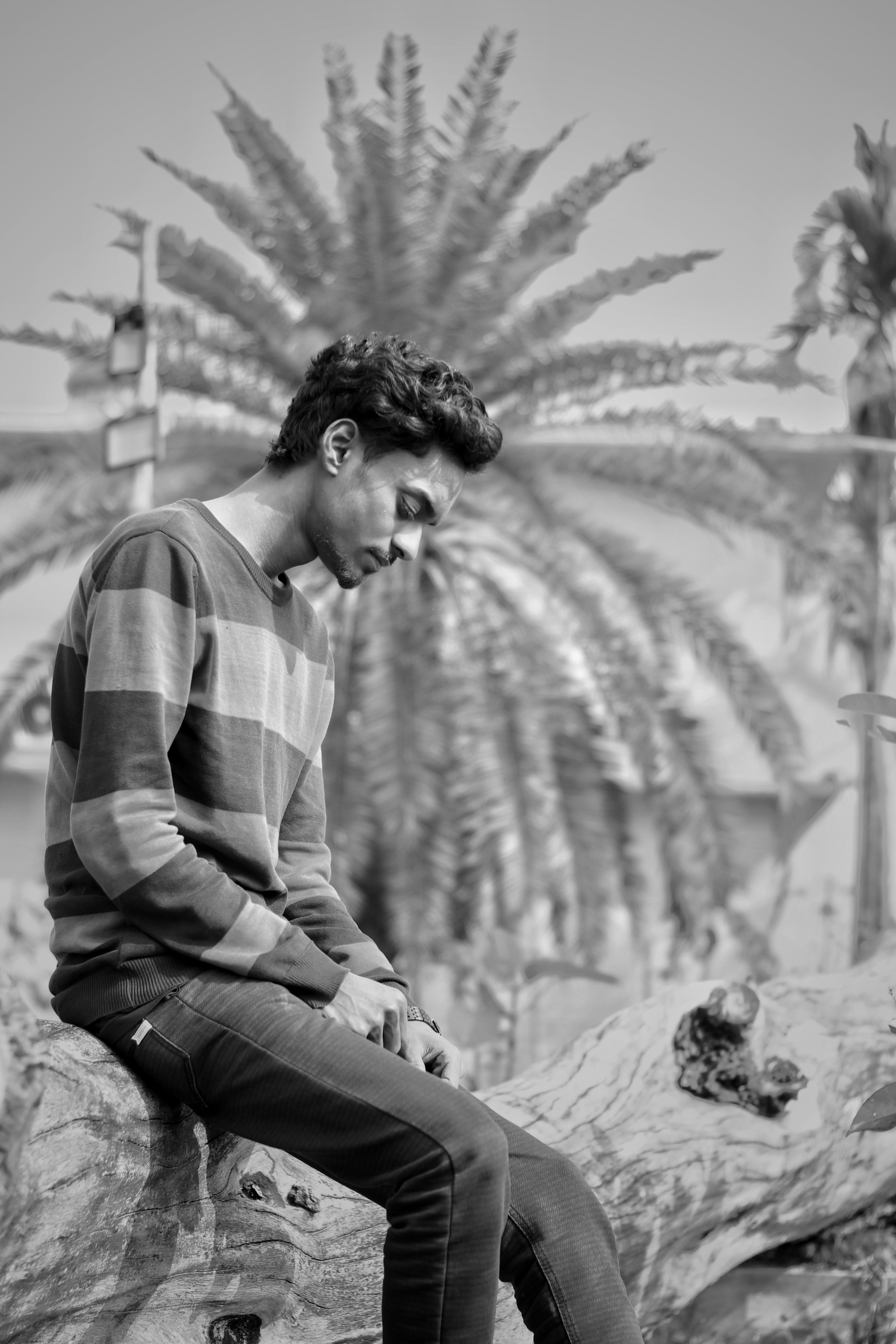 Black and white photo of a young man sitting on a tree trunk outdoors, reflecting.