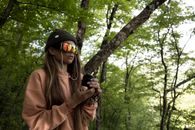 Woman in Brown Coat Wearing Black Knit Cap Taking Photo of Green Trees