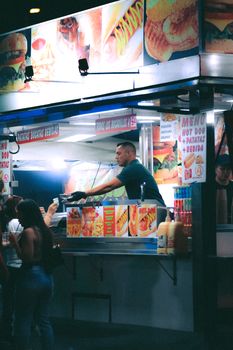 Busy street food vendor serving customers at night market stall with bright lights and menu signs.