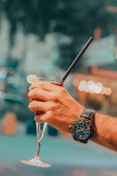 A hand holding a cocktail glass with a visible wristwatch against a blurred nightlife backdrop.