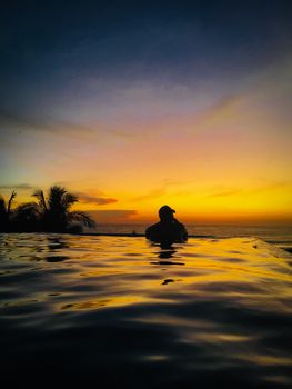 A silhouette in a luxurious infinity pool at sunset with palm trees and vibrant sky.