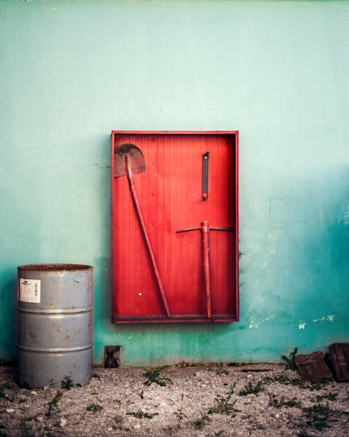 Red Wooden Shelf with Shovel and Axe Beside a Rusty Steel Drum