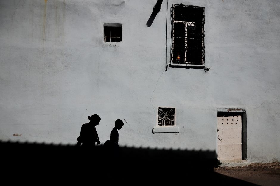 Silhouettes of two people walking against a rustic urban wall illuminated by shadows.