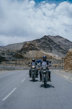 Two motorcyclists ride through a scenic mountainous road under a cloudy sky.