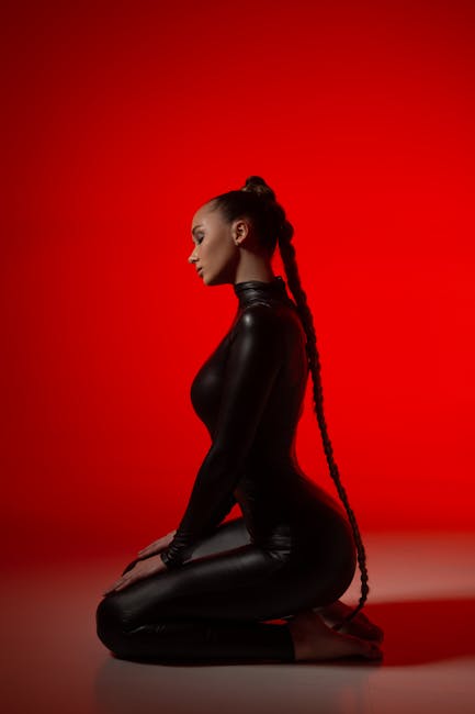 Profile of a kneeling woman in a sleek black bodysuit with a long braid, set against a vibrant red backdrop.