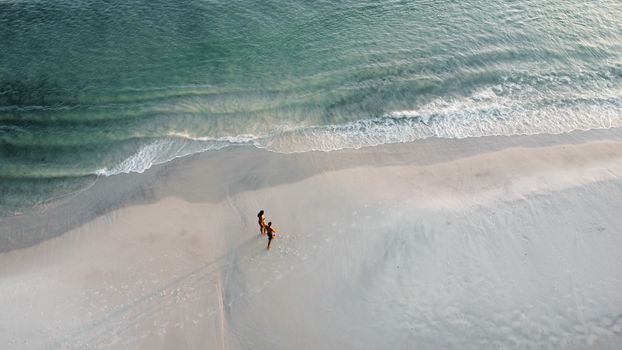Peaceful aerial scene of a couple walking on a serene oceanfront beach with gentle waves.
