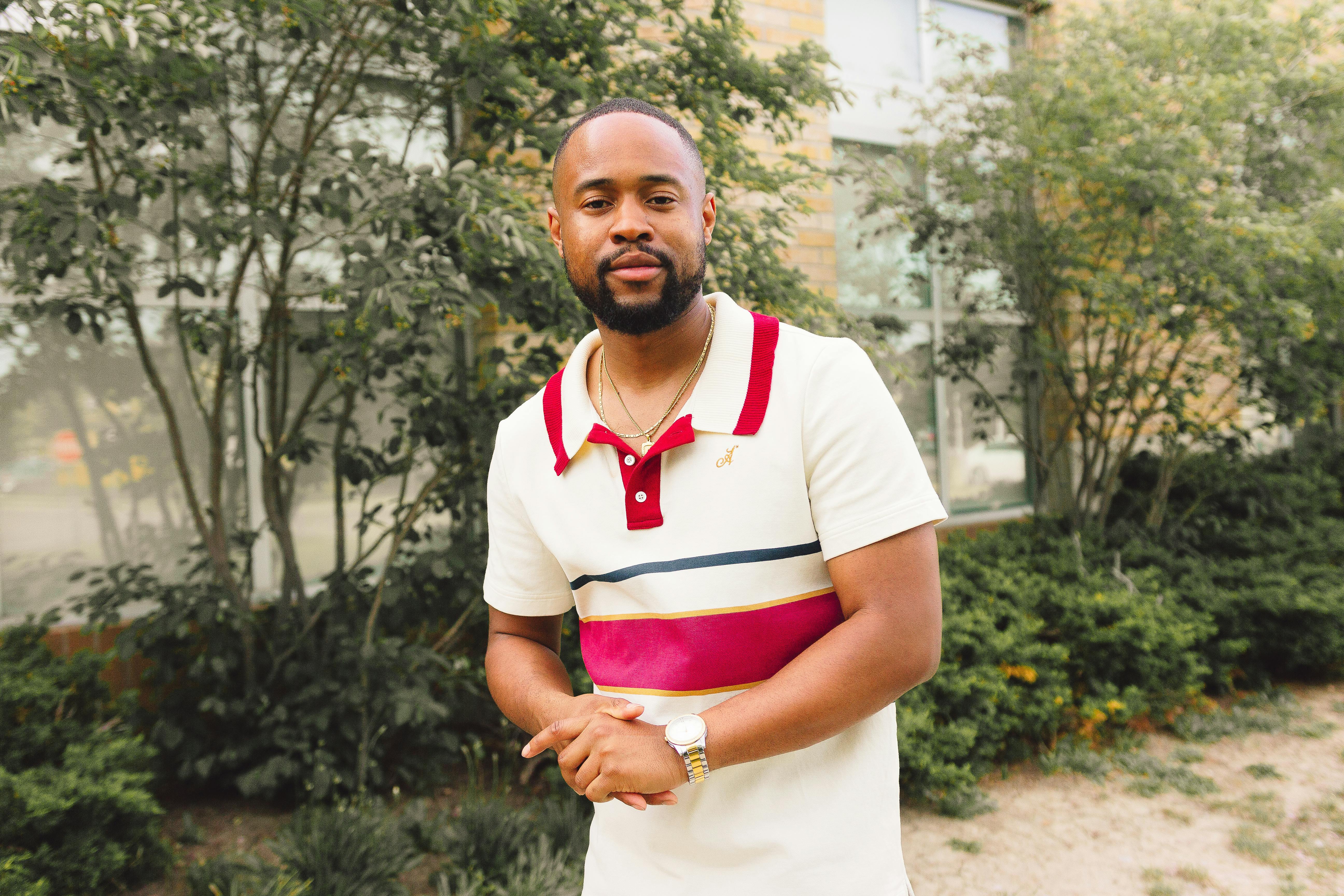 Confident young man in stylish polo shirt posing outdoors with greenery around.