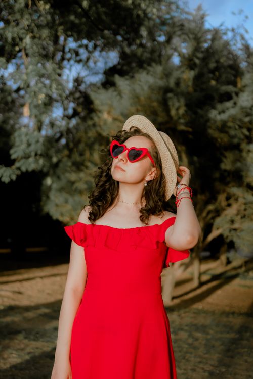 Free A fashionable woman outdoors in a red off-shoulder dress, wearing heart-shaped sunglasses and a sun hat. Stock Photo