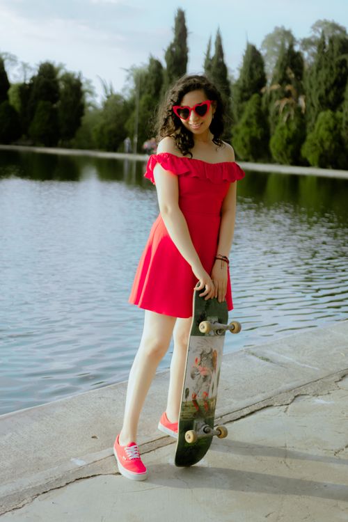 Free Stylish woman in red dress and sunglasses posing with skateboard by a serene lake. Stock Photo