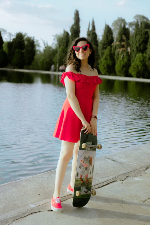 Free Stylish woman in red dress holding skateboard by a serene lake in a park setting. Stock Photo