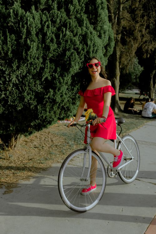 Free A smiling woman rides a bicycle in a park, wearing a vibrant red dress and sunglasses. Stock Photo