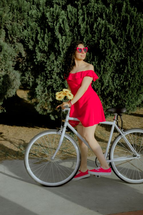 Free A stylish woman in a red dress stands beside a bicycle in a sunny park. Stock Photo