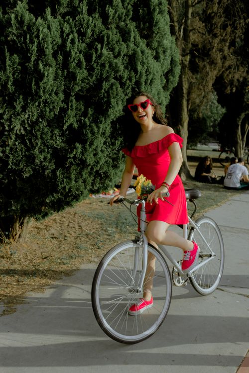 Free Woman in red dress and sunglasses joyfully rides a bicycle through a sunny park path. Stock Photo
