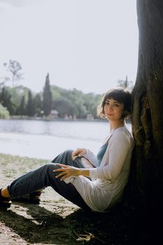 A serene portrait of a woman sitting by a tree near a lake, enjoying the outdoors.