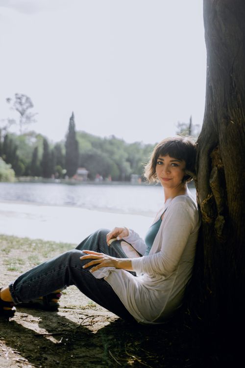 Free A serene moment of a woman sitting by a tree near a river in a sunny park. Stock Photo