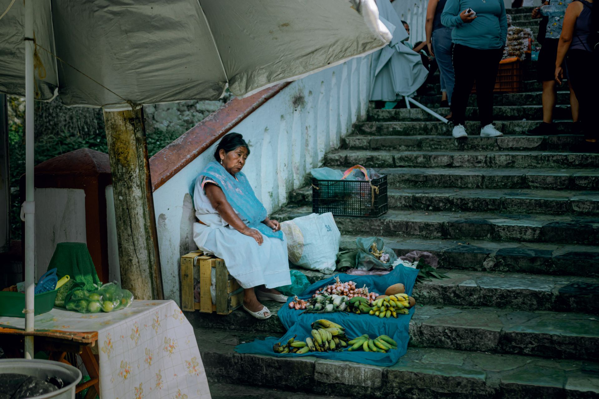 Outdoor market scene with a vendor selling fruits while seated on stone steps in a bustling area.