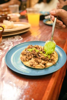 A close-up of a taco with meat and green salsa being prepared on a blue plate at a dining table.
