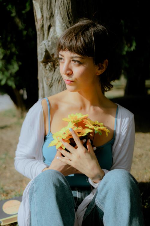 Free A young woman in a serene park holding vibrant yellow flowers, basking in sunlight. Stock Photo