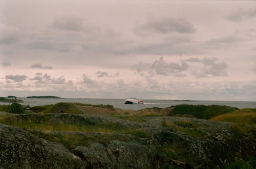 Free Photo of a Grey Cloudy Sky Above a Rocky Hill Partially Covered with Grass and a Boat Sailing in a Sea  Stock Photo