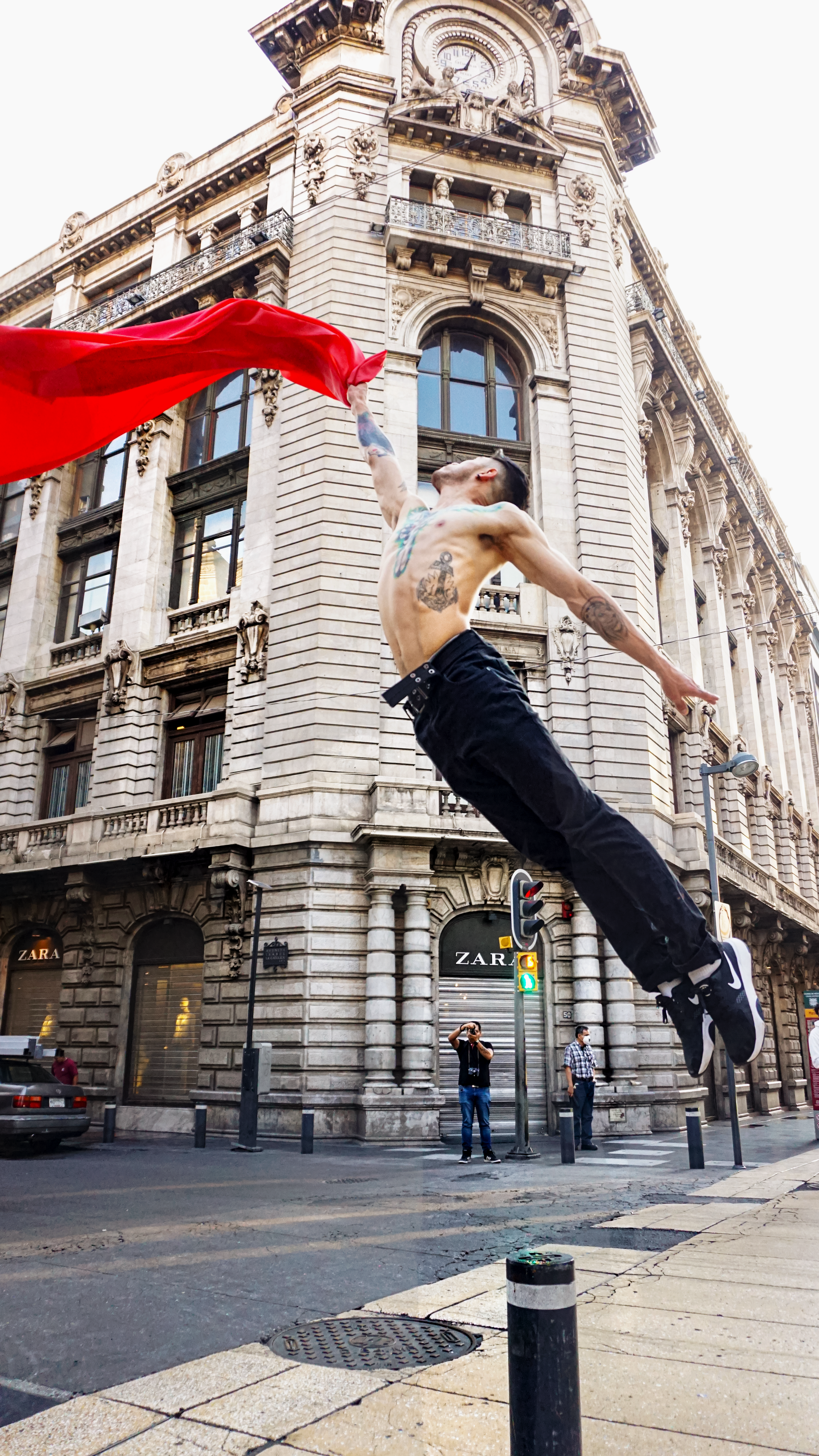 A shirtless man with tattoos jumps gracefully in front of historic architecture in Mexico City.