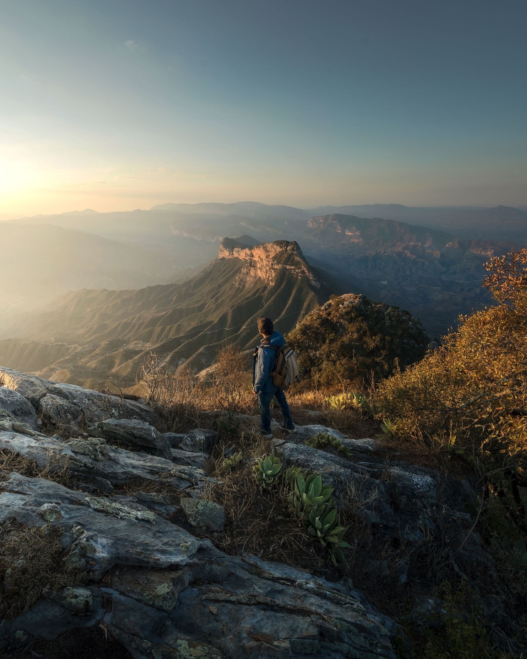 A hiker with a backpack standing on a rocky outcrop above a vast canyon, soft low sunlight washing over the ridges