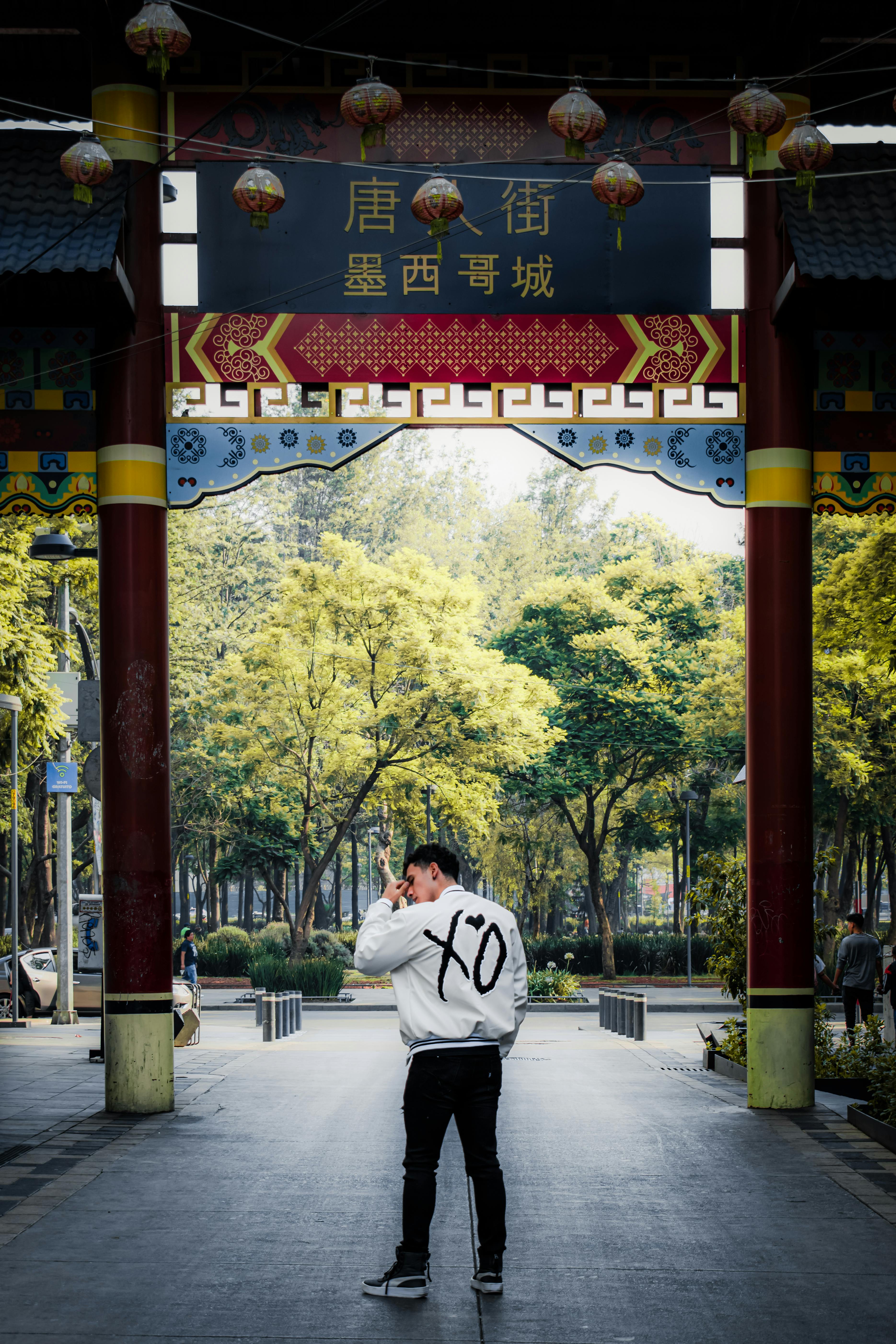 Man with XO jacket walking under colorful Chinese archway surrounded by trees.