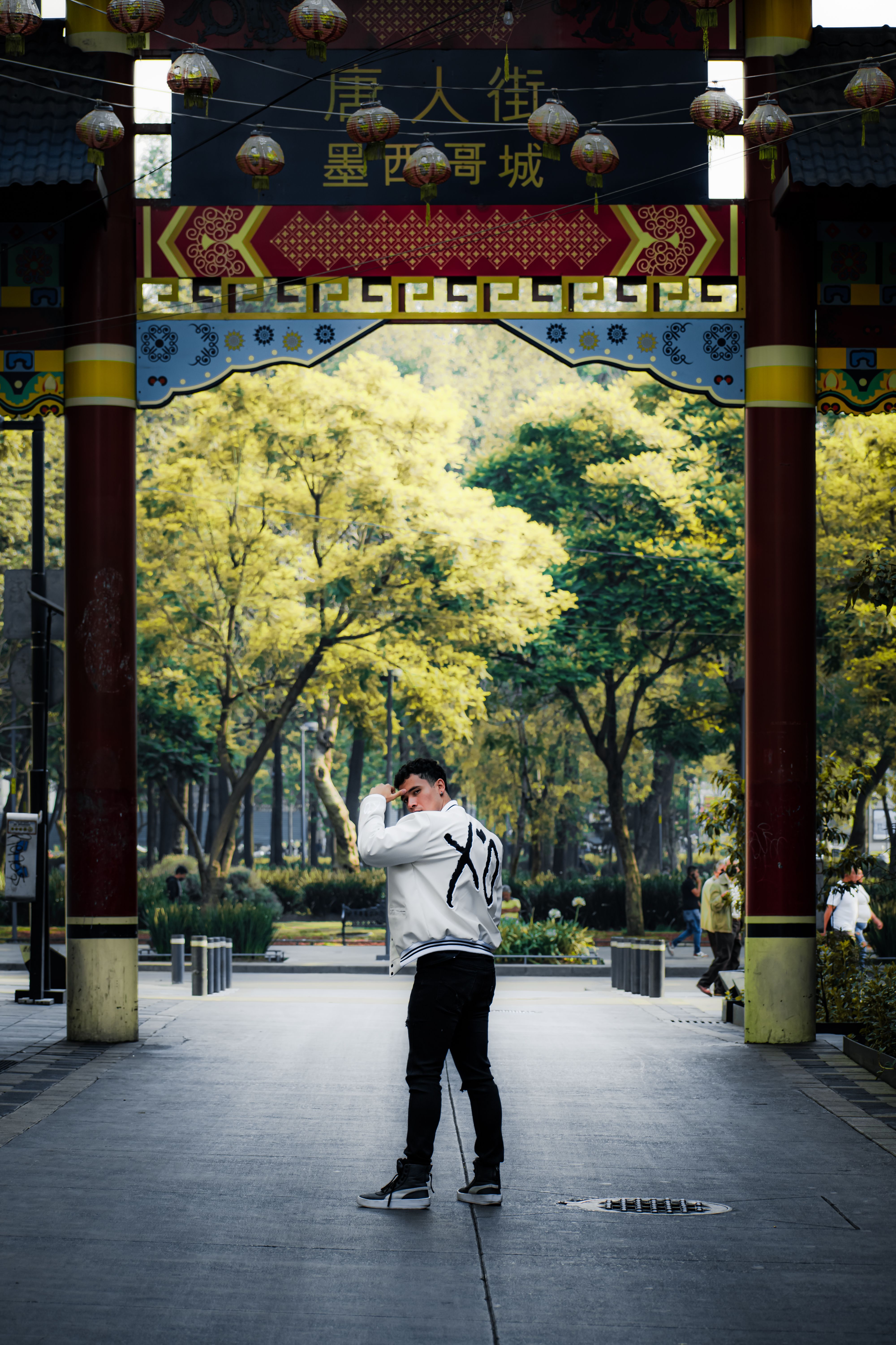 A person in a white jacket poses under a decorative Chinese archway in a lush park.