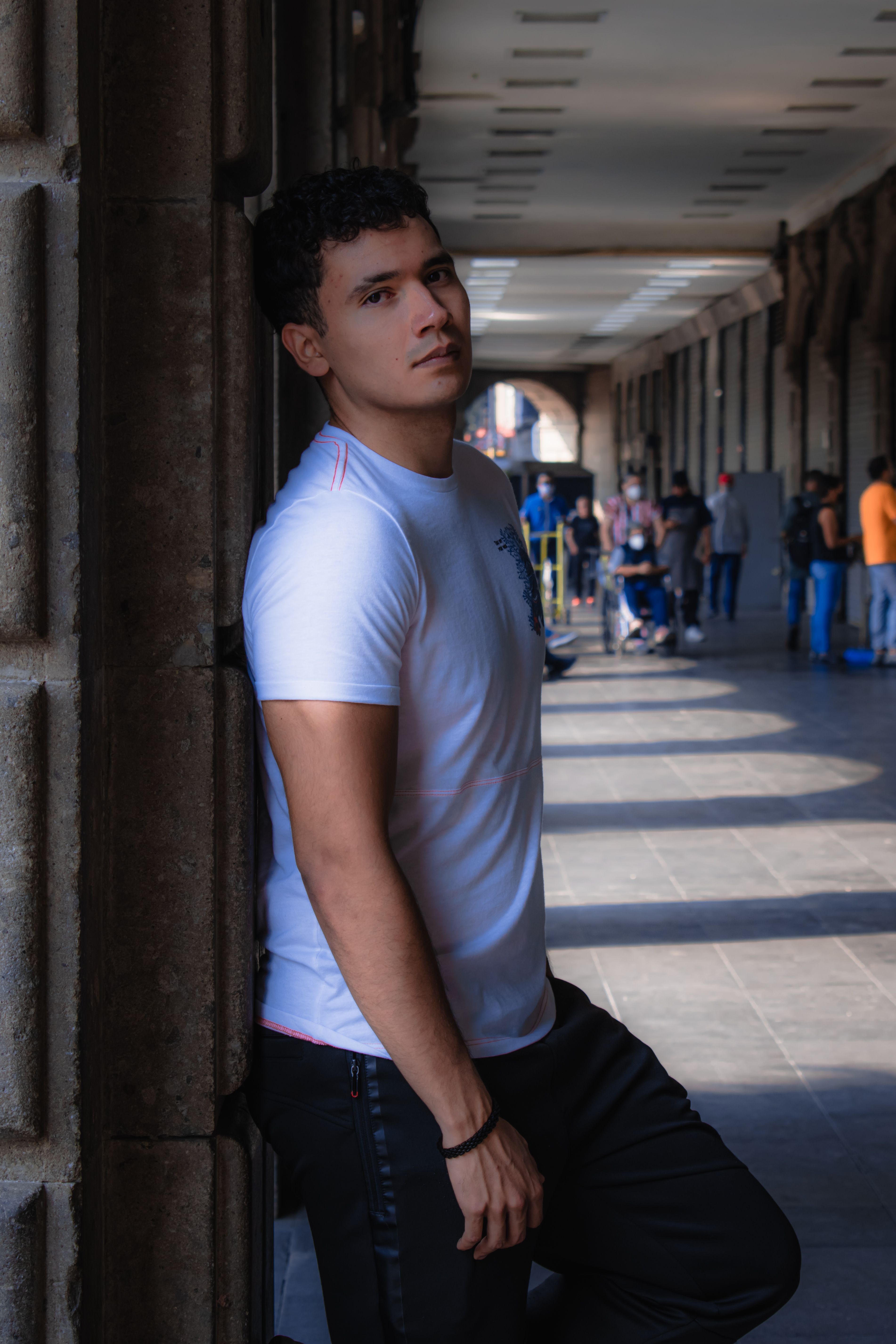 Portrait of a young man in a white shirt leaning against a wall in an outdoor corridor.