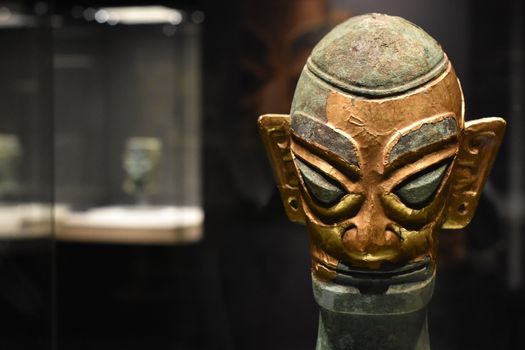 Close-up of an ancient bronze mask exhibited in a dimly lit museum display case.
