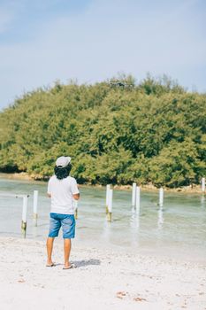 Man operating a drone on a sandy beach with lush greenery in the background on a sunny day.