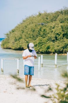 A man in casual summer attire flies a drone on a sunny beach, surrounded by clear waters and lush greenery.
