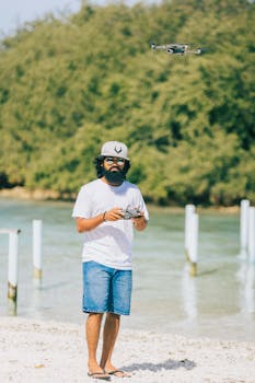 A man with facial hair and a hat operates a drone on a sunny beach with clear water and trees.