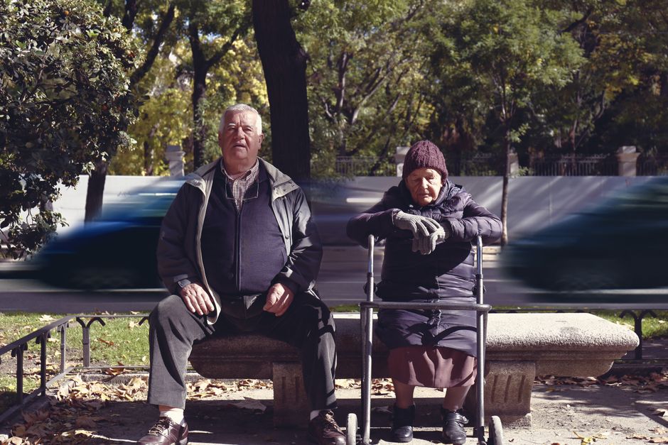 Pareja de ancianos disfrutando de un día soleado en un banco en el parque de Madrid, con coches borrosos pasando.