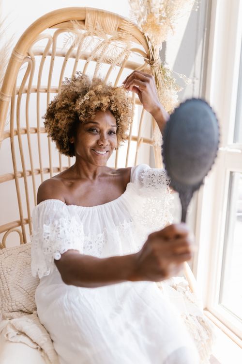 Free A woman in a white dress sits in a wicker chair, smiling as she looks at her reflection in a mirror. Stock Photo