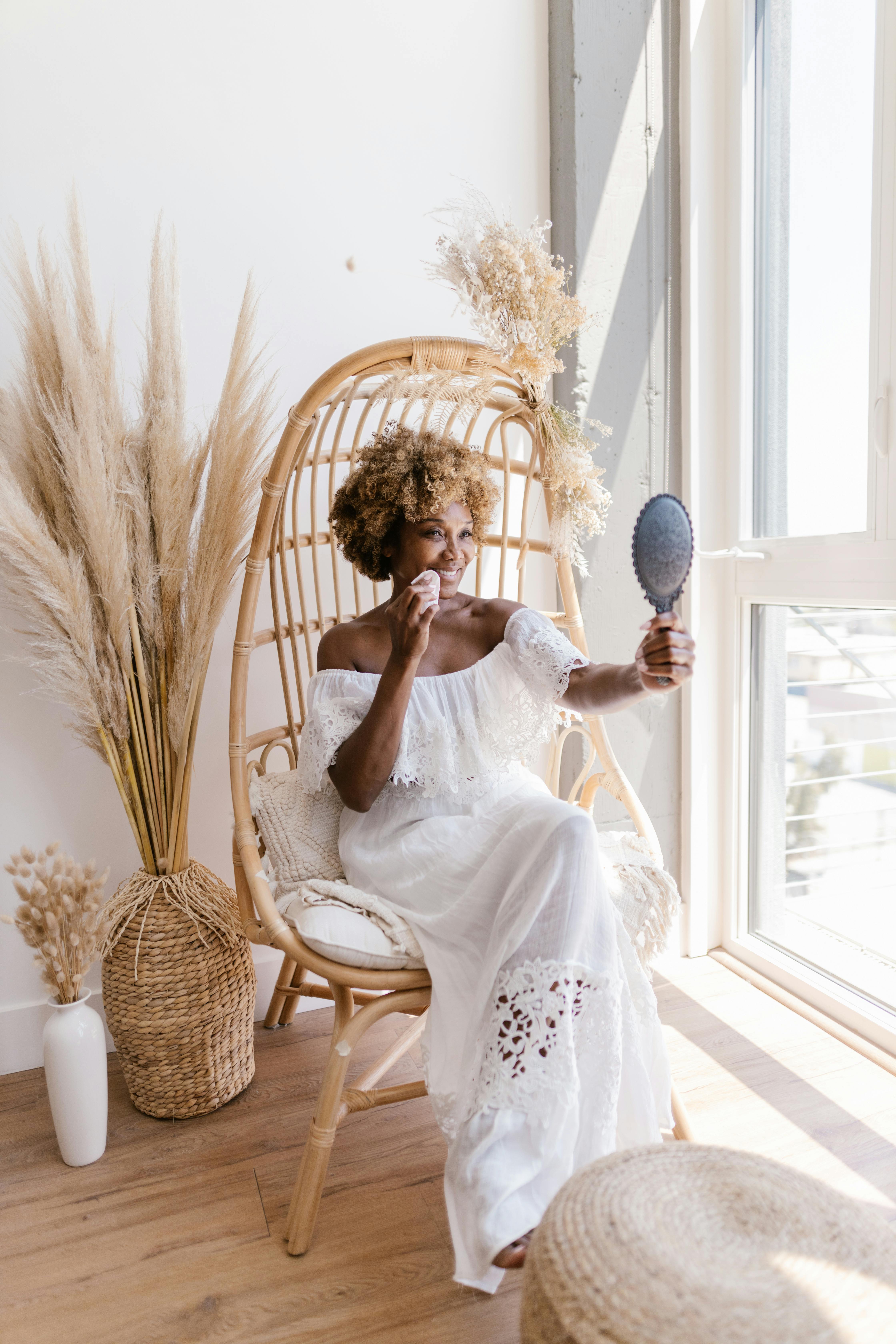 Free A stylish woman in a white dress admires herself in a mirror, seated in a wicker chair beside a window. Stock Photo