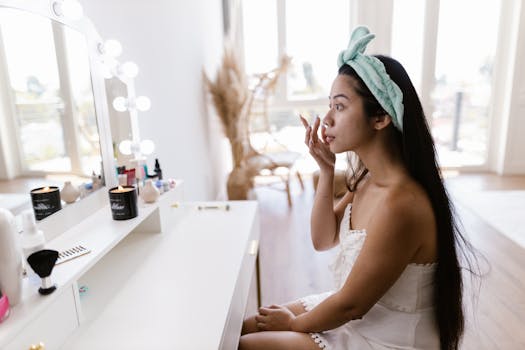 A woman applying makeup at a vanity mirror with a headband, candles, and beauty products.