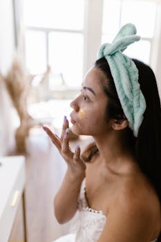 Woman in a towel applies skincare cream to her face, wearing a headband, in a well-lit room.