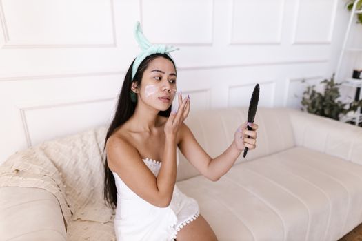 Asian woman in a towel applying face cream while looking in a handheld mirror indoors.