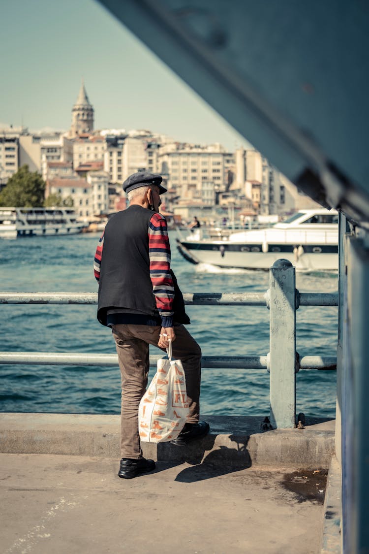 Photo Of An Elderly Man Standing Next To A Railing Looking At The Harbour And A Boat Sailing Against The Background Of City Buildings