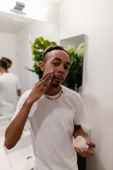 Man applying face cream in a bathroom mirror while wearing a white t-shirt, focusing on skincare routine.