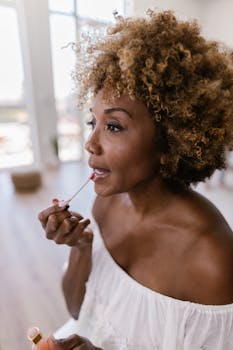 An adult woman applies lip gloss by a window, in a bright and airy indoor setting.