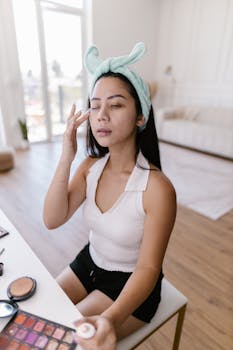 Young woman in casual attire applying makeup indoors with natural light.