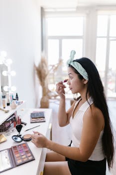 A young woman applies makeup at an illuminated vanity table indoors.