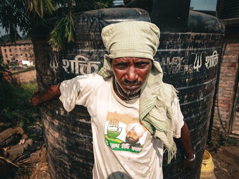 A man with traditional headwear stands against a large tank, showcasing rural life.